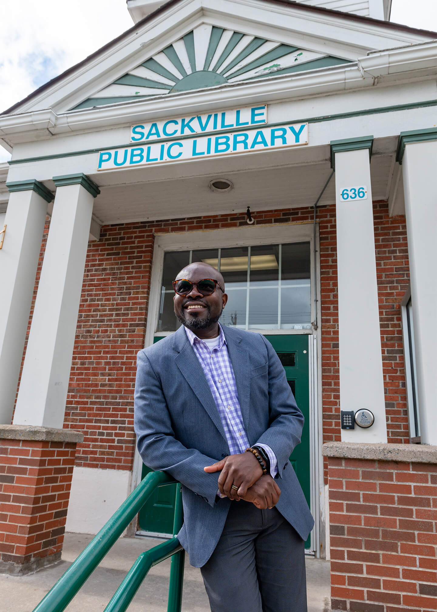 Ayo is wearing a blue suit and sunglasses, leaning on the railing in front of the Sackville Public Library entrance.